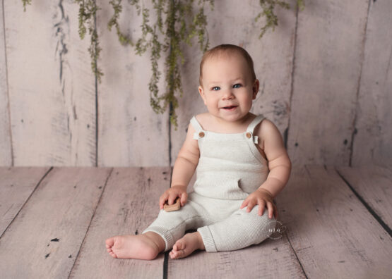 metro Detroit baby photographer Melissa Anne Photography one year old boy with light gray knit overalls on wood floor smiling