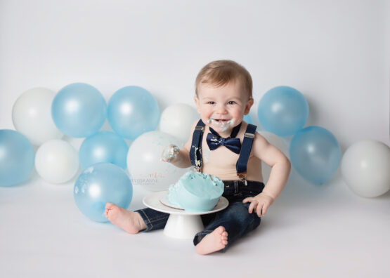 Troy Michigan Cake Smash Melissa Anne Photography first birthday photo shoot with boy wearing jeans with suspenders and bowtie with blue balloons eating cake and smiling